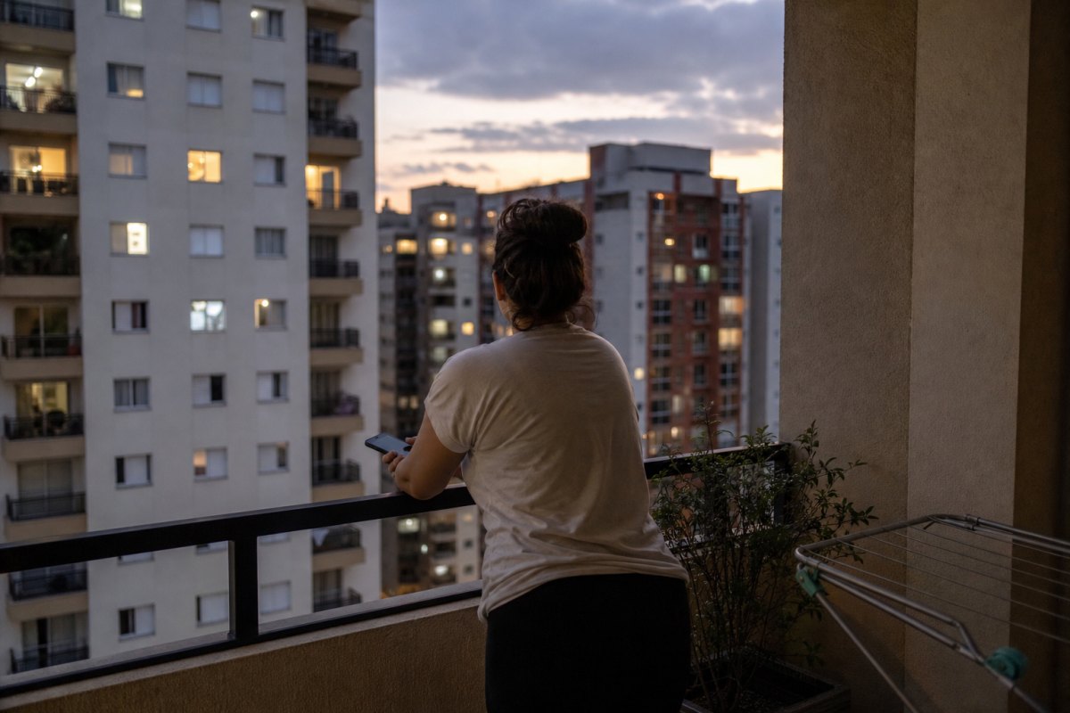 Moradora de costas na varanda do apartamento ao entardecer, com celular na mão e prédios vizinhos iluminados